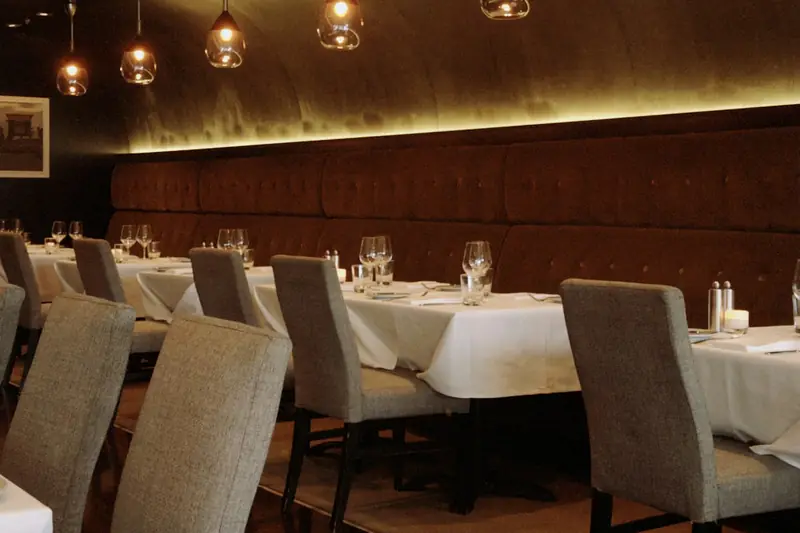 Restaurant dining room with a padded banquette, laid white tables, upholstered chairs and pendant lights