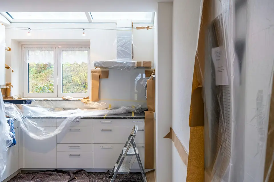 Bright kitchen mid-renovation, with white cabinets covered in protective plastic and paper, a stepladder and a window